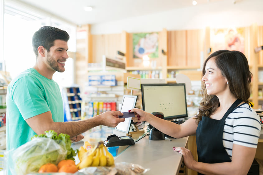 Cliente sorridente passando o cartão para atendente em um caixa de supermercado, com frutas e legumes sobre o balcão. Palavra-chave: atendimento ao cliente.
