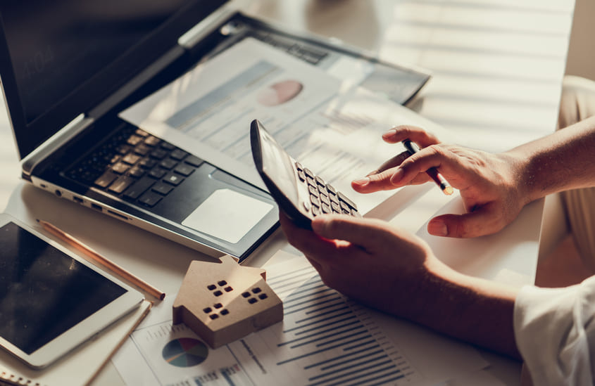 Pessoa calculando finanças com calculadora e documentos fiscais ao lado de um notebook e miniatura de casa.