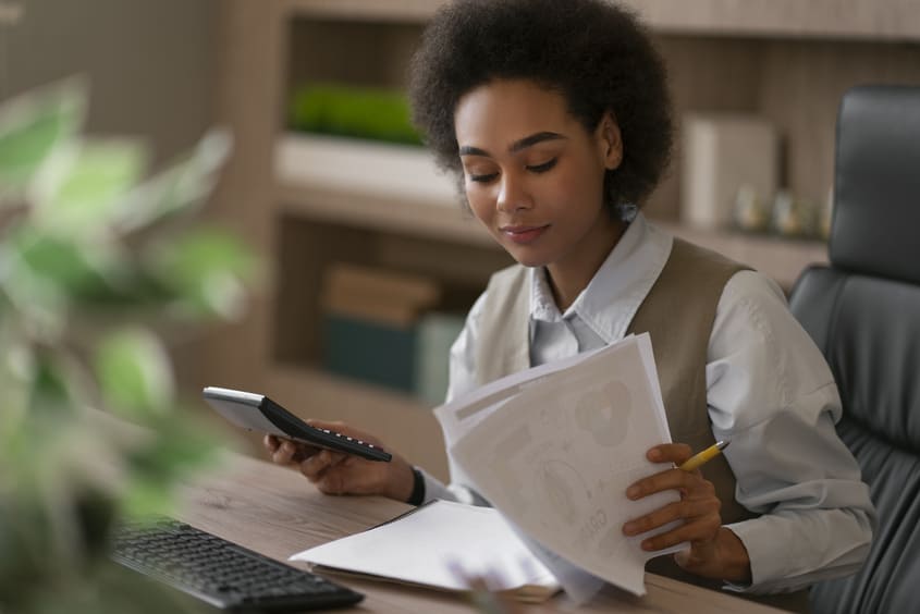 Mulher analisa documentos e utiliza calculadora em escritório, lidando com obrigações fiscais.