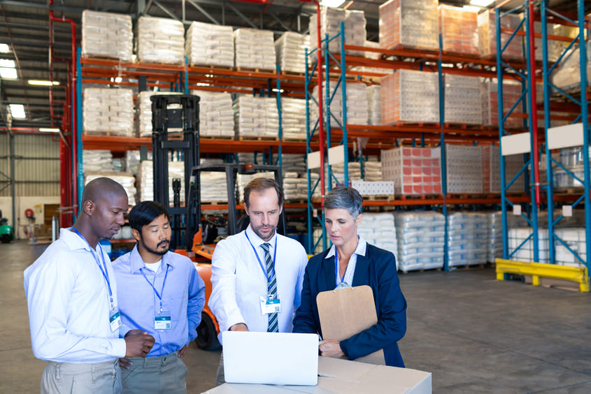 Profissionais realizando conferência de mercadorias com notebook em um armazém logístico, com prateleiras altas repletas de produtos embalados ao fundo.