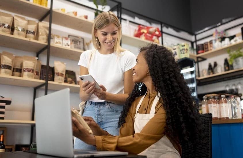 Duas mulheres interagindo em uma loja moderna com prateleiras ao fundo, uma segurando um celular e a outra sentada com um notebook.