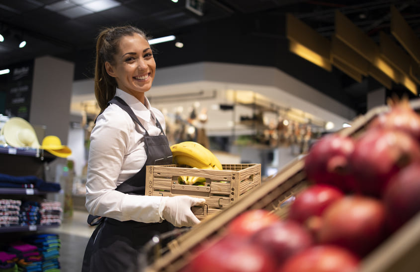 Funcionária sorridente em supermercado segurando caixa de bananas, com frutas em destaque ao fundo, ideal para campanhas sobre como fazer promoções no varejo.