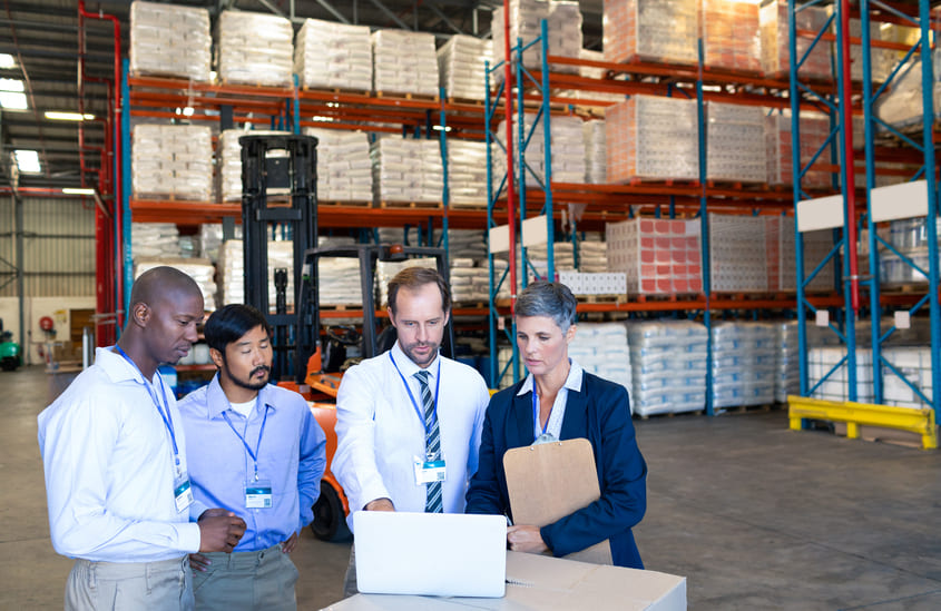 Equipe de profissionais realizando conferência de mercadorias em armazém logístico, analisando dados em um notebook.