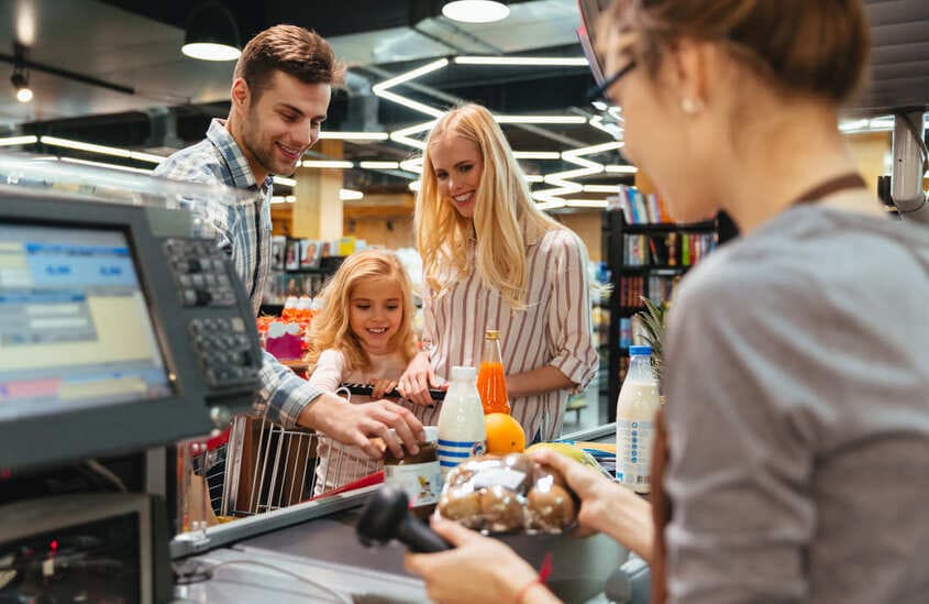 Família sorridente realiza pagamento no caixa de um supermercado, enquanto a atendente passa os produtos.