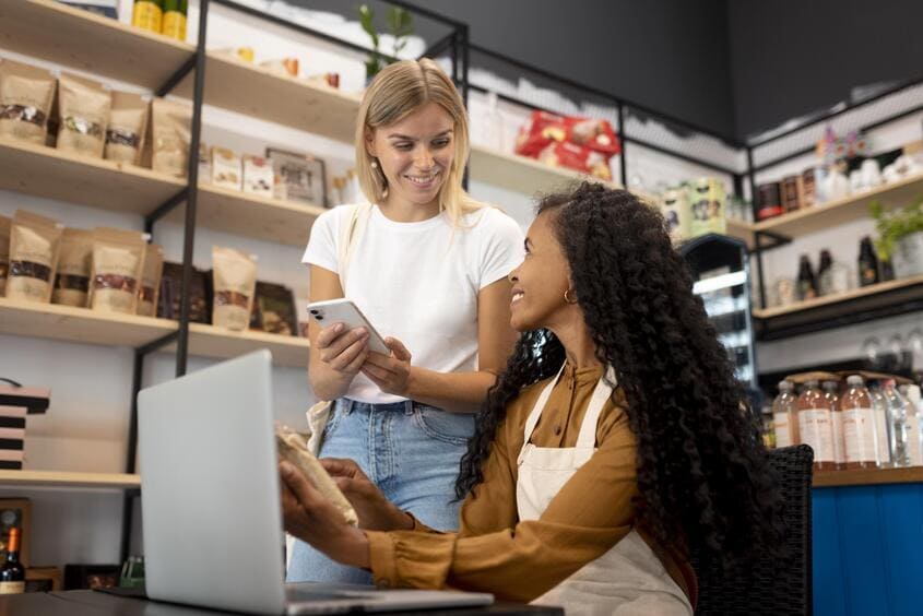 Duas mulheres conversam em uma loja, uma segurando um celular e a outra usando um notebook, com prateleiras de produtos ao fundo.