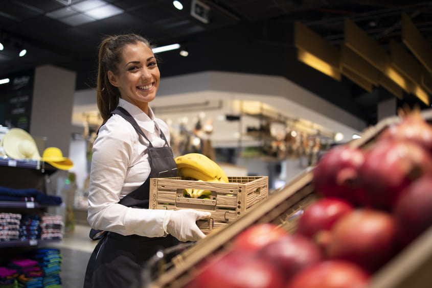 Funcionária sorridente de supermercado segurando uma caixa de bananas, em frente a prateleira de frutas com maçãs vermelhas em destaque.