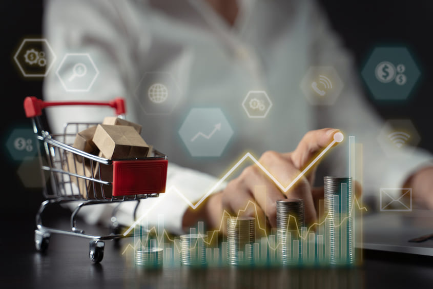 Carrinho de compras em miniatura com caixas de papelão e pilhas de moedas em destaque, com gráficos de crescimento e ícones digitais ao fundo, simbolizando estratégias de vendas e crescimento financeiro.