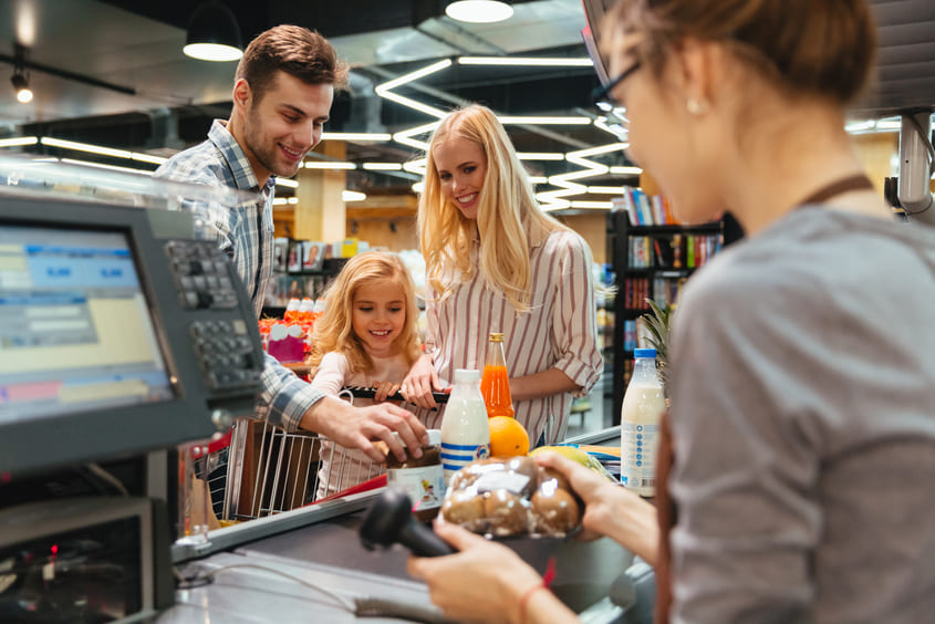 Família sorridente no caixa de supermercado, com pai, mãe e filha observando a operadora registrando as compras, incluindo leite, refrigerante e frutas.